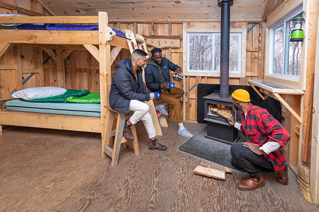Three people inside a cabin with a wood stove and bunk beds.