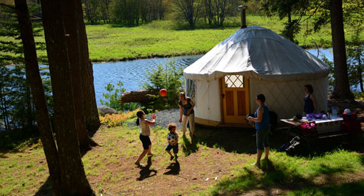 A wood and canvas yurt surrounded by trees on the shore next to a river.