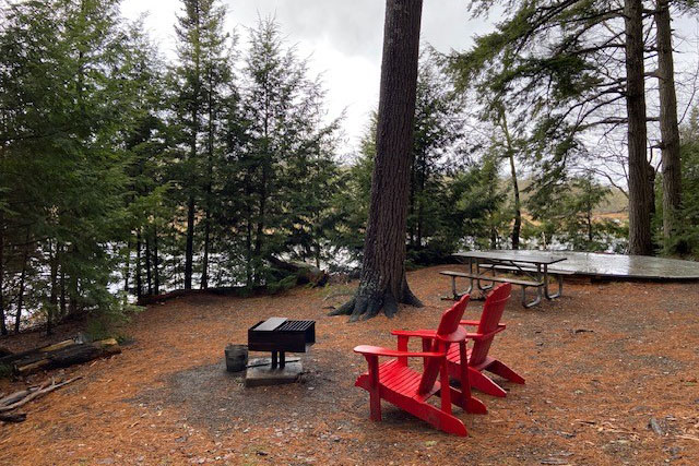 A tent site with a round tent pad, picnic table, fire box, and two red chairs surrounded by trees, next to the river.