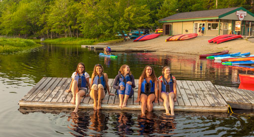 Five young people wearing life jackets and sitting on the dock with canoes and kayaks on the shore in the background.