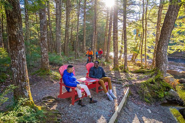 Two visitors sit in the Parks Canada red chairs beside the trail with two cyclists in the background.