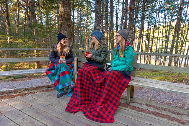 Three visitors sit on a bench with cups of coffee and blankets.