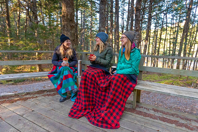 Trois visiteurs assis sur un banc avec des tasses de café et des couvertures.