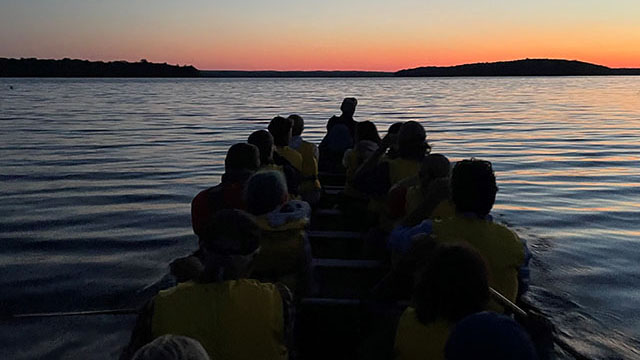 A group of people in a big canoe at sunset.