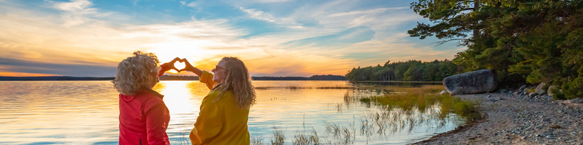 Un coucher de soleil au bord de l’eau et deux femmes formant un cœur avec leurs mains. 