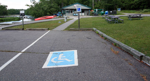 The accessible parking spot at Jakes Landing with canoe rental building, canoes, docks, picnic tables and grass in the background.
