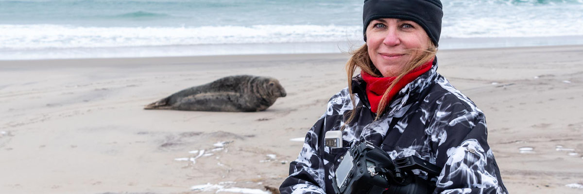 A visitor with a camera on the beach with a seal in the background.