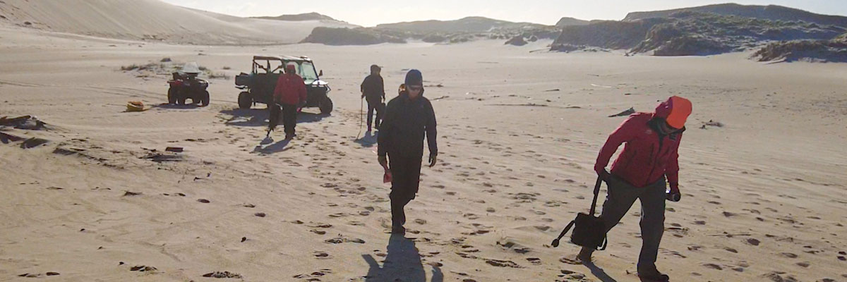 Four people walk across the sand from two all-terrain vehicles with sand dunes in the background.