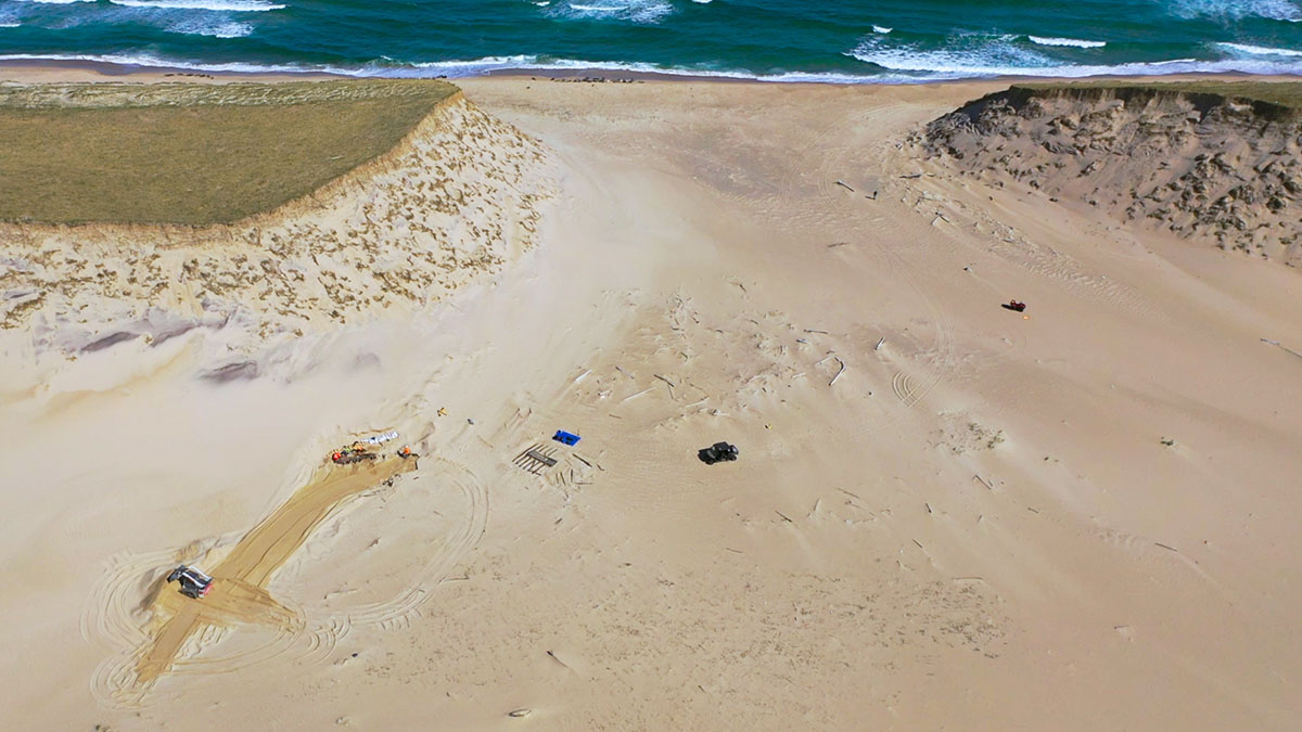 An aerial view of the dig site showing the site is in an area open to the water between two tall sand dunes.