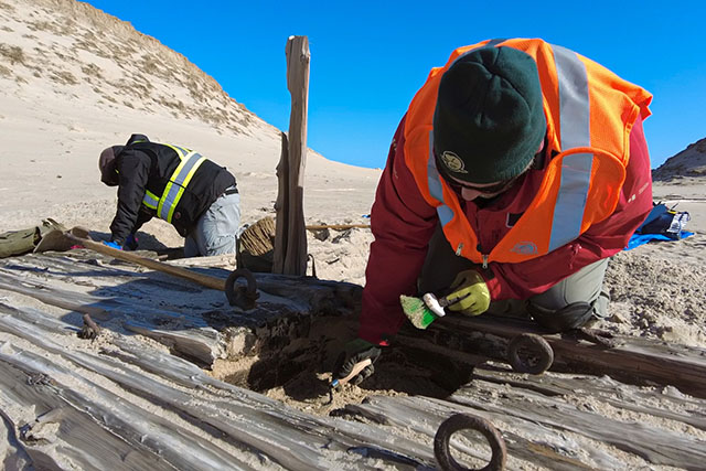 Two people kneel on the ground and use hand tools to remove sand from an exposed section of the wreck.
