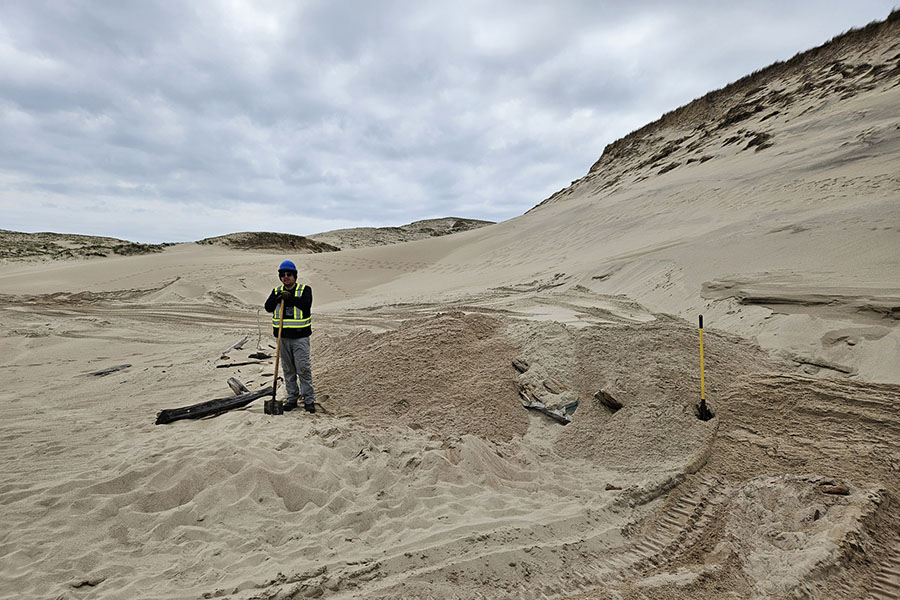 A person stands holding a shovel next to a pile of sand, where the wreck section is buried.