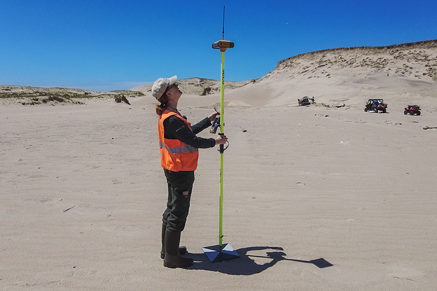 A person balances an 8-10 ft pole with an antennae on the sand; a small datalogger is attached to the middle.