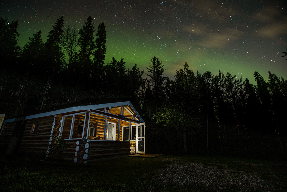 Image taken at night of a brown log cabin. Green Northern Lights appear along the treetops and stars pepper the night sky.
