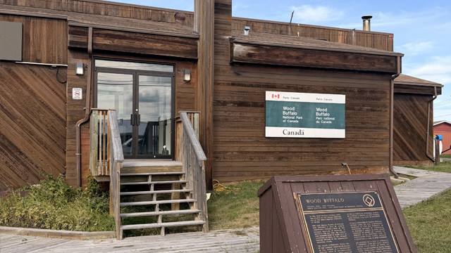 Exterior view of the Wood Buffalo National Park Visitor Centre in Fort Chipewyan, Alberta. The wooden building features Parks Canada signage, a small staircase leading to the entrance, a boardwalk in the foreground, and a commemorative plaque on the lawn under a clear blue sky.