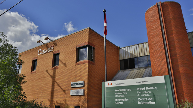 Exterior view of the Wood Buffalo National Park Visitor Centre in Fort Smith, Northwest Territories. Under a blue sky with scattered clouds, the red brick building features Parks Canada signage. In the foreground, there is a Canadian flag on a flagpole, a large bilingual information centre sign and a green sandwich board with opening hour information.