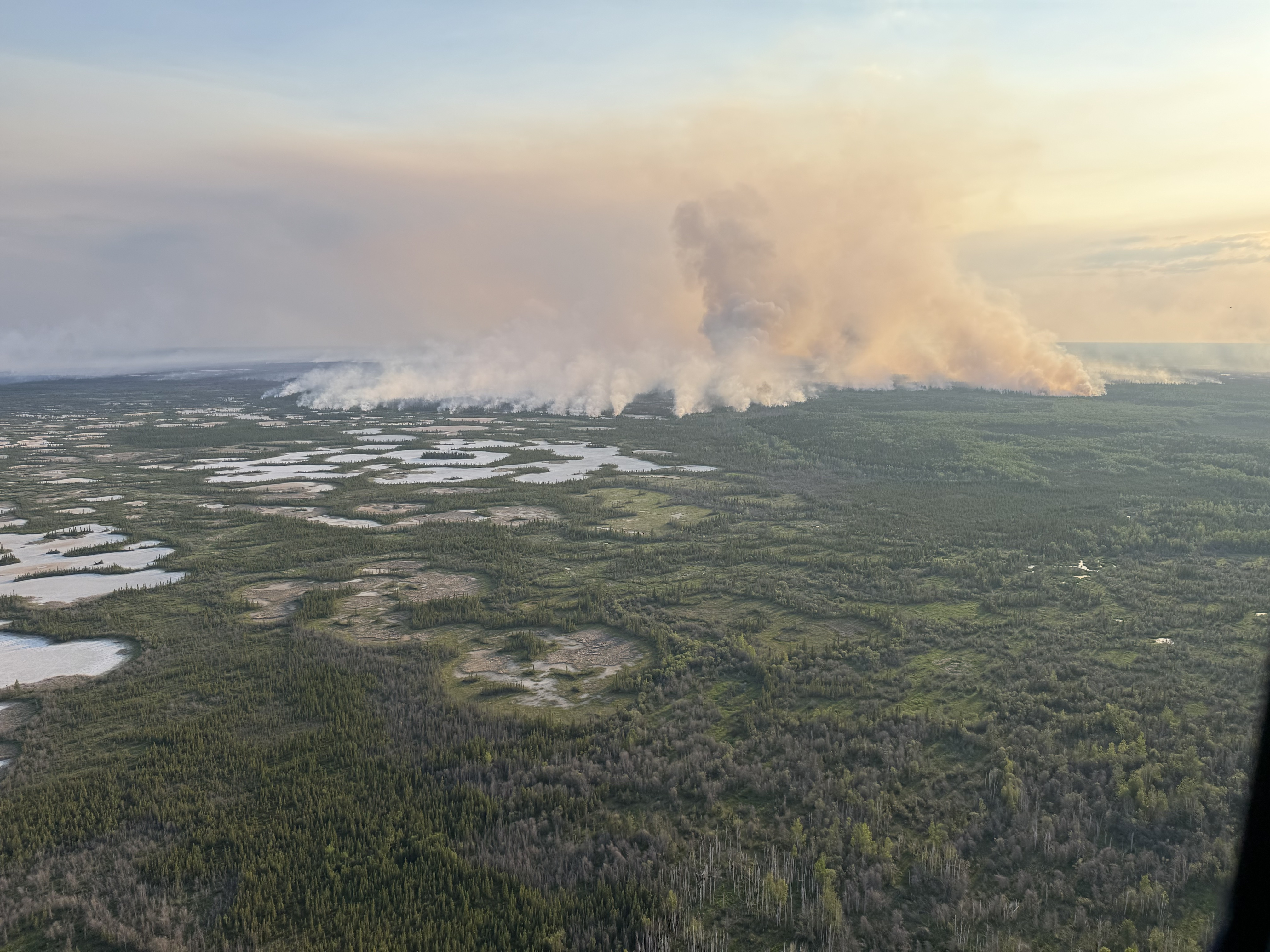 Une vue aérienne montre un grand feu de forêt qui brûle dans un paysage boisé parsemé de zones d’eau. Des colonnes de fumée s’élèvent dans le ciel et se répandent à l’horizon.