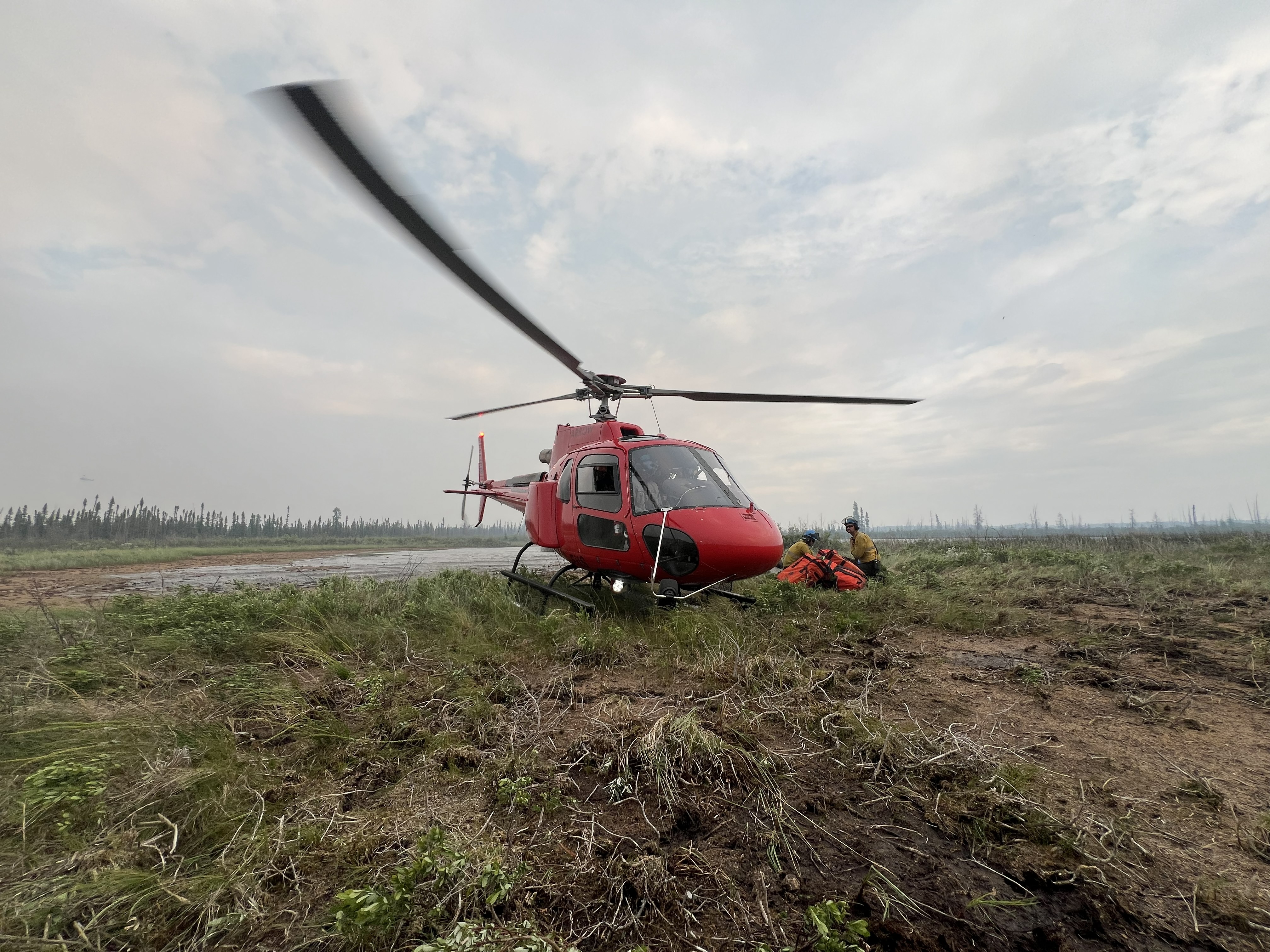 Un hélicoptère rouge est stationné sur une zone dégagée tandis que le personnel de lutte contre les incendies de forêt s’agenouille en toute sécurité à proximité, avec les pales du rotor de l’avion floues en mouvement sur un ciel nuageux et un paysage récemment brûlé.