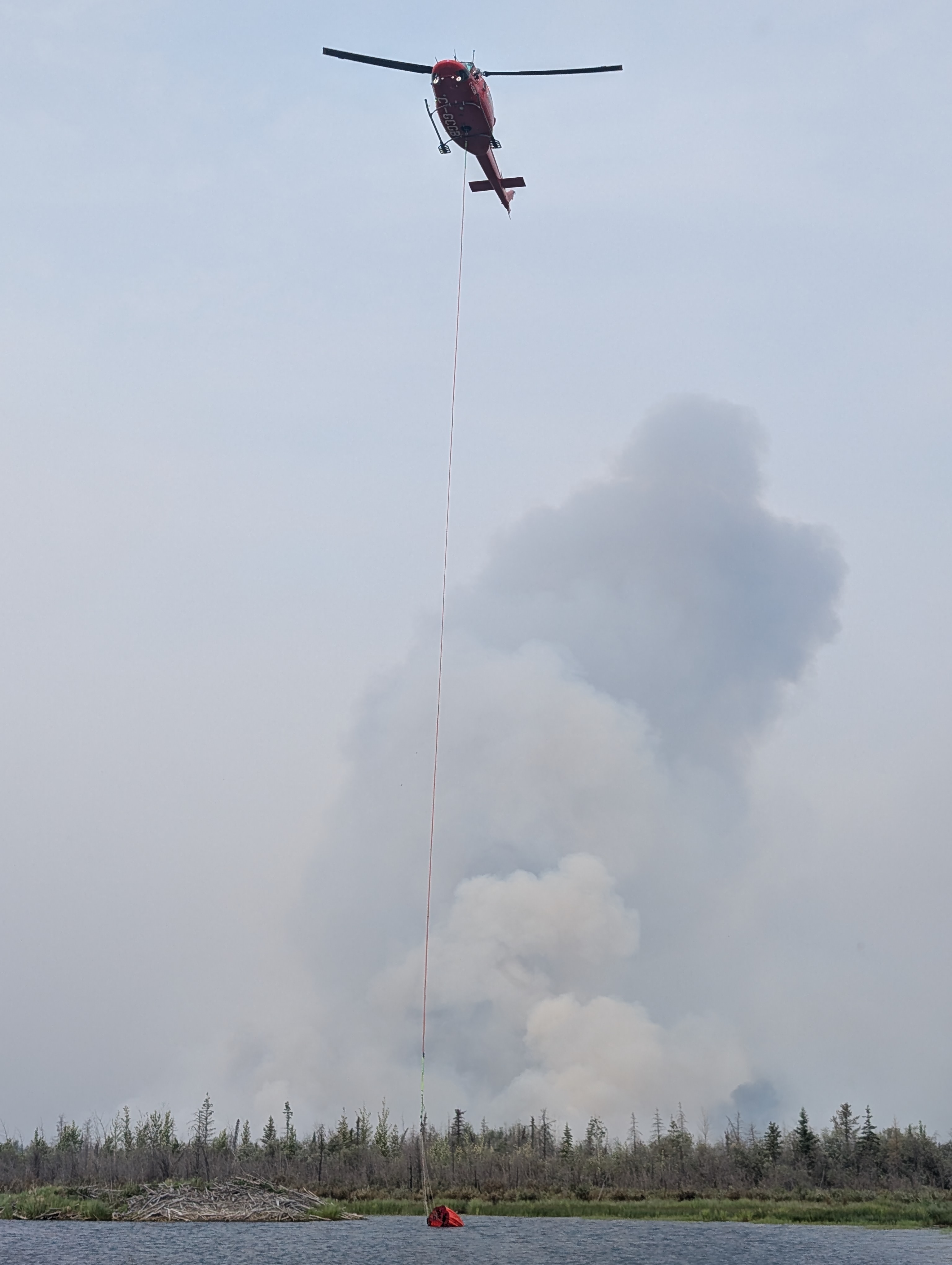 Un hélicoptère plane avec une longue ligne de seau abaissée vers un plan d’eau tandis que de grandes colonnes de fumée s’élèvent de la forêt en feu en arrière-plan.