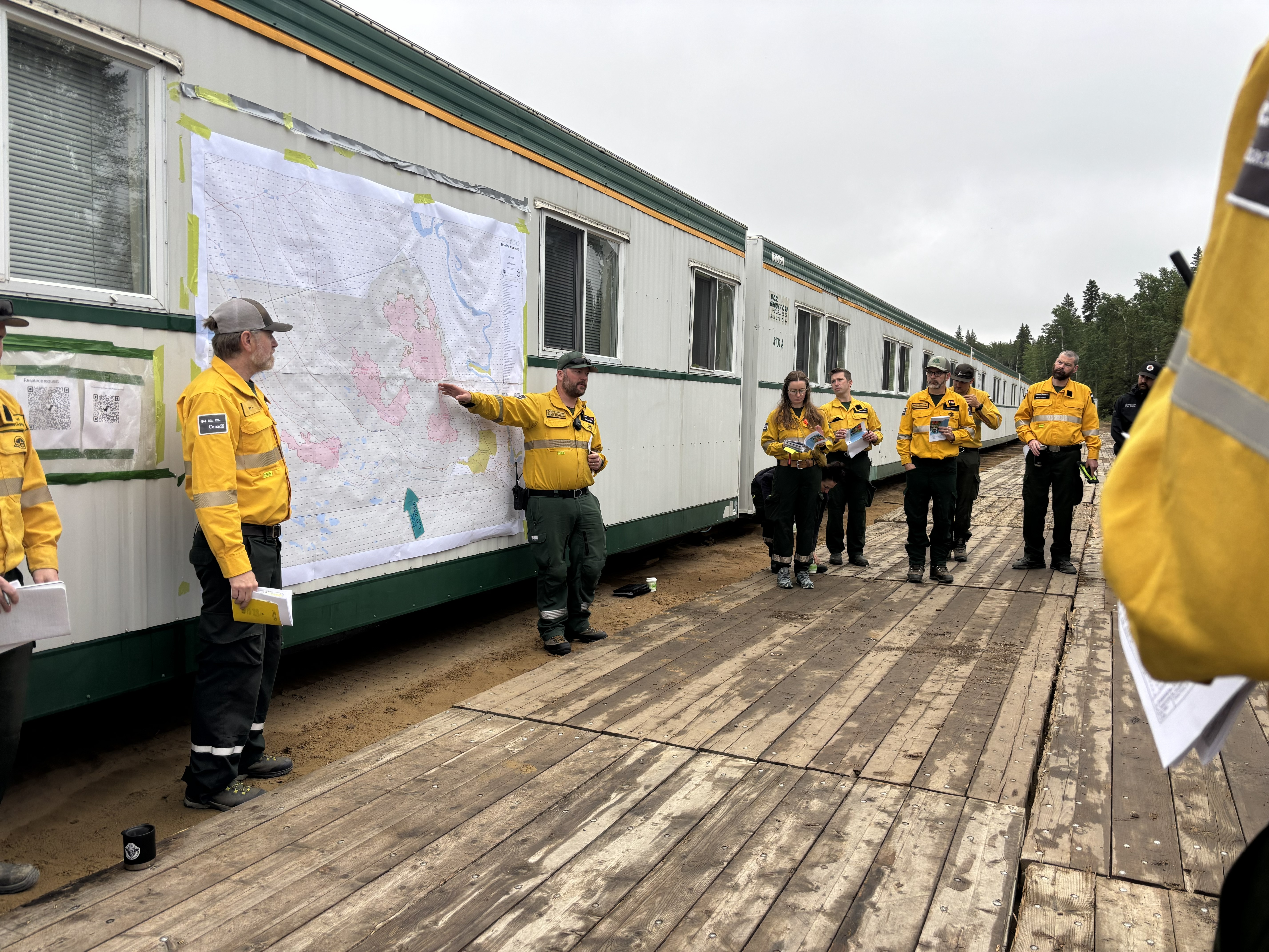 Des employés de lutte contre les incendies de forêt en Nomex jaune se tiennent sur une terrasse en bois à côté d’une grande carte murale, participant à une séance d’information opérationnelle à l’extérieur d’une roulotte de camp portative.