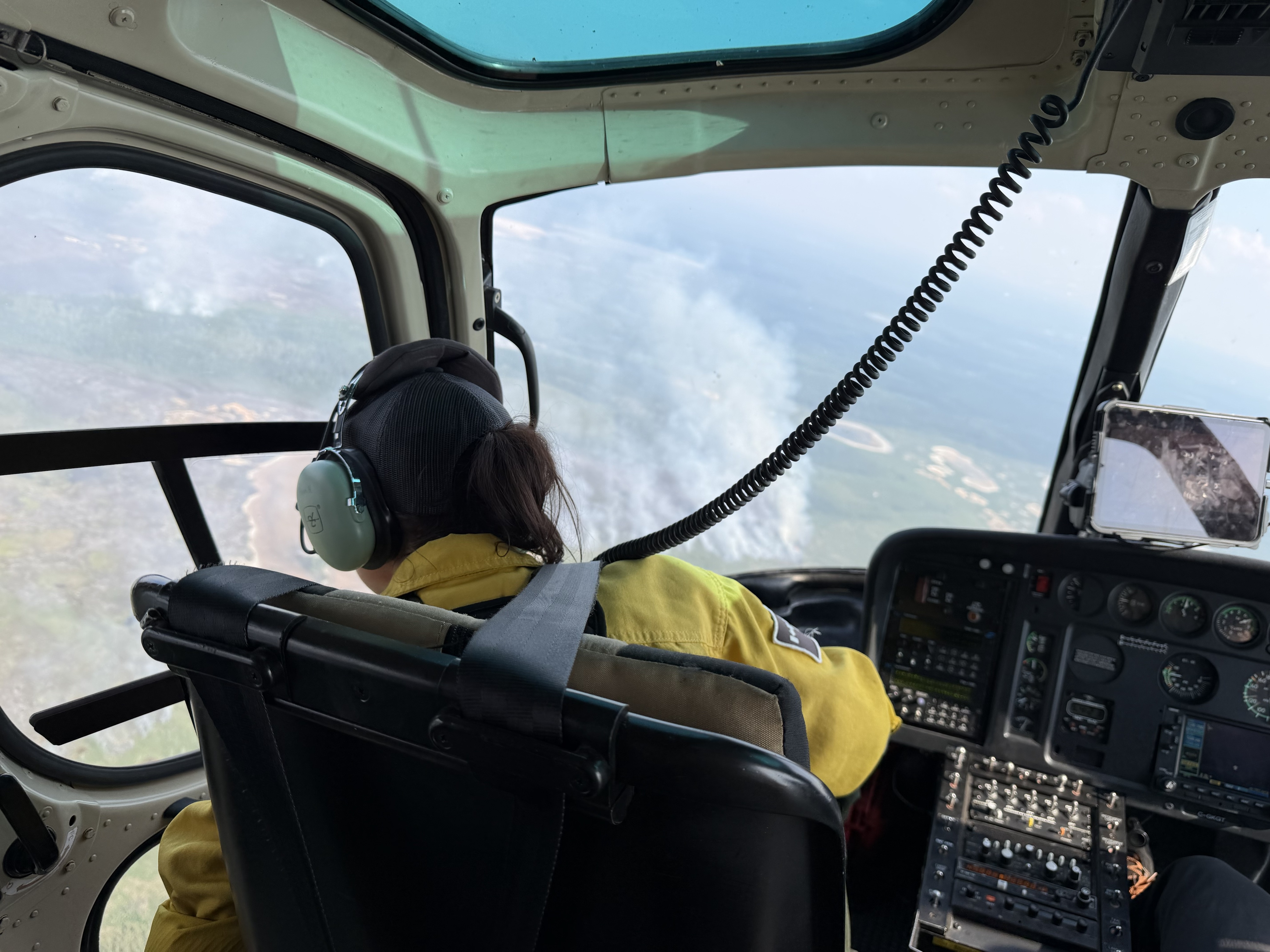 Un pompier forestier vêtu d’un Nomex jaune et d’un casque d’écoute est assis dans le cockpit d’un hélicoptère, regardant à travers le pare-brise un paysage rempli de fumée lors d’une opération aérienne de lutte contre les incendies de forêt.
