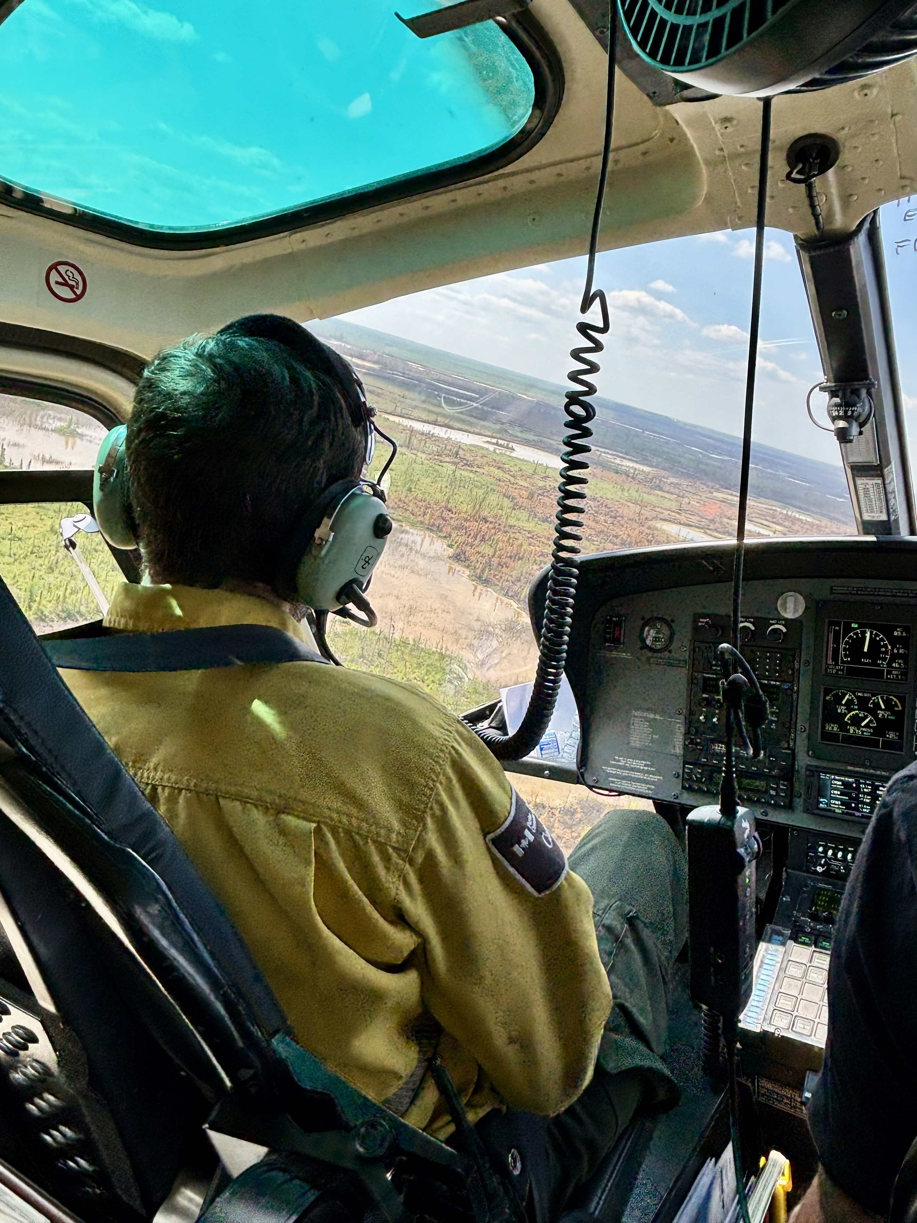 Un spécialiste portant un Nomex jaune et un casque d’écoute est assis dans le cockpit d’un hélicoptère, surveillant le paysage en contrebas lors d’une opération aérienne de feu de forêt.