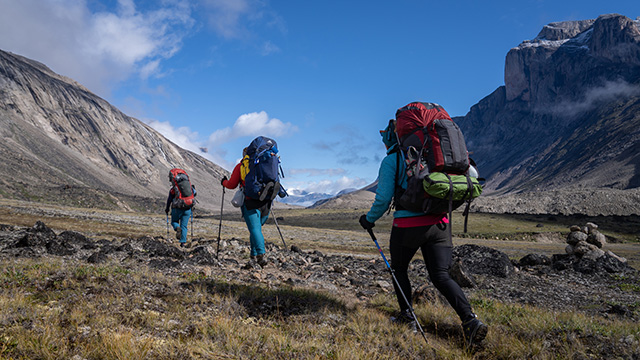 Trois randonneurs avec des sacs à dos traversant la toundra et entourés de montagnes. 