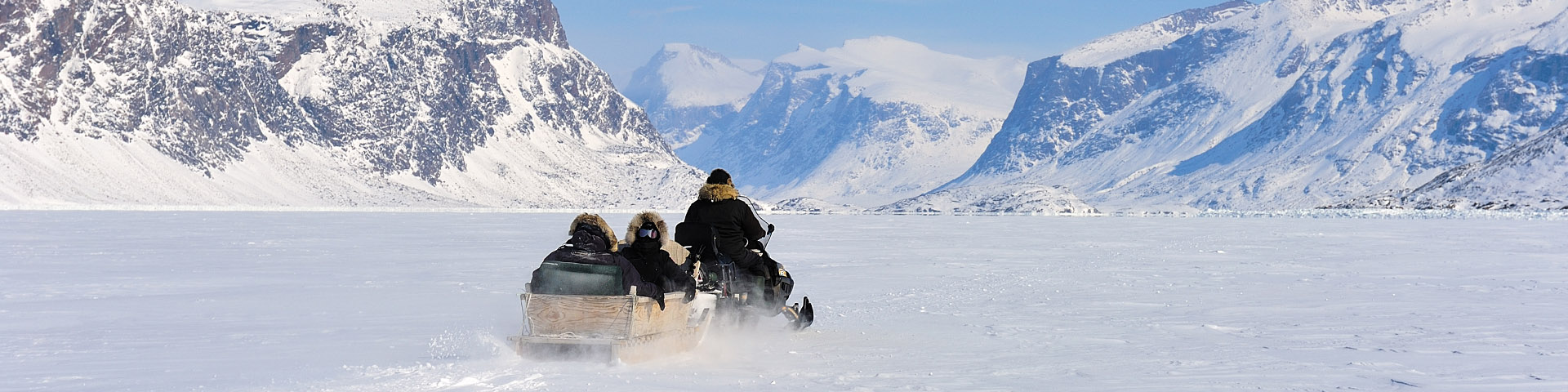 Un skidoo tirant deux personnes dans un qamutik à travers la glace de mer avec un paysage de montagne en arrière-plan.