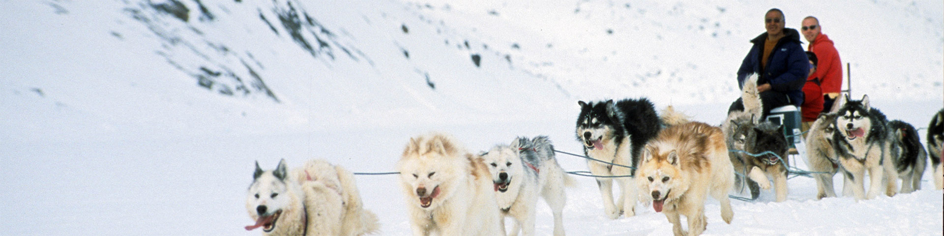 Une équipe de chiens de traîneau tirant trois personnes sur un qamutik à travers un paysage enneigé.