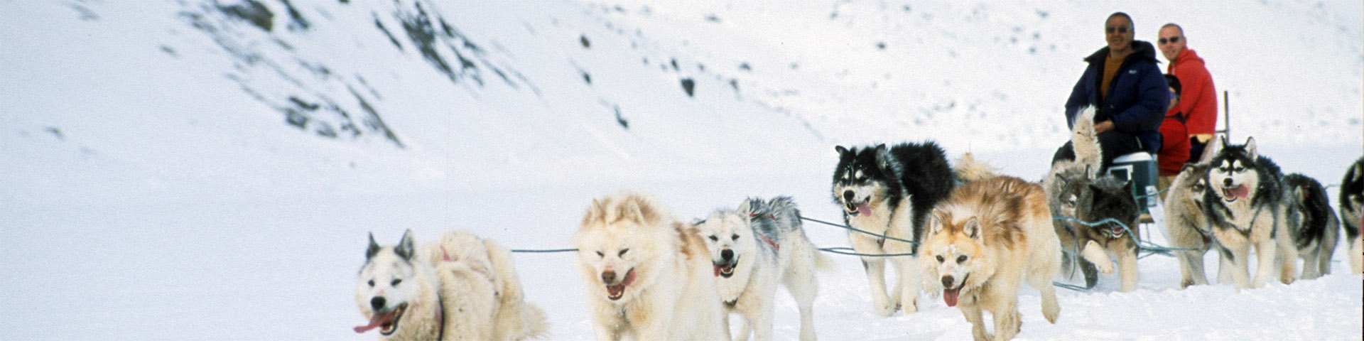 A dogsled team pulling three people on a qamutik through a snowy landscape.