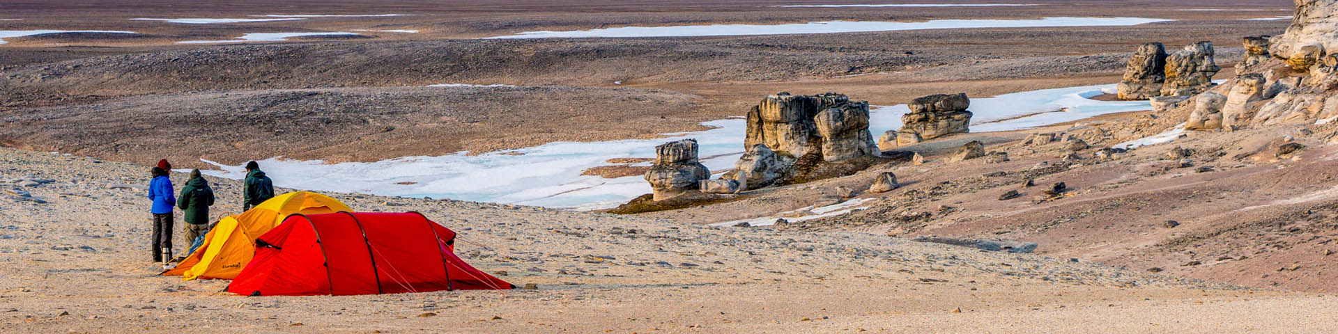 People camping in a wide-open, rocky plain with patches of snow, surrounded by rugged terrain.