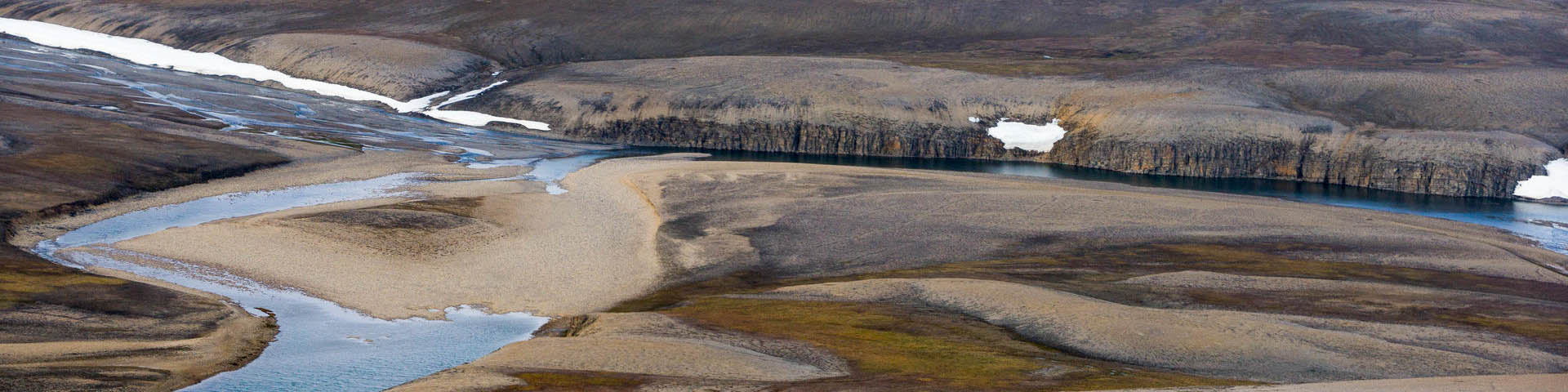 A bird's-eye view of a wide-open tundra plain with patches of snow and scattered vegetation.