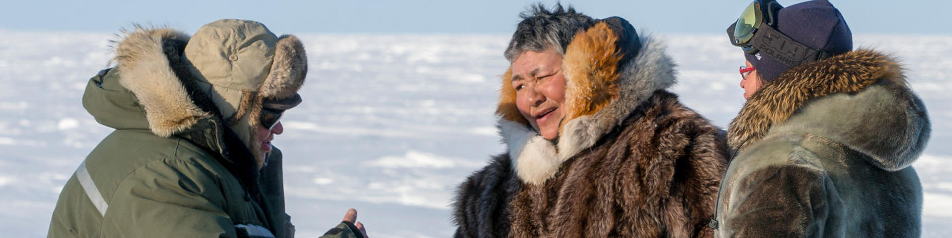 Three people in heavy winter coats and gear, talking on a snowy landscape.