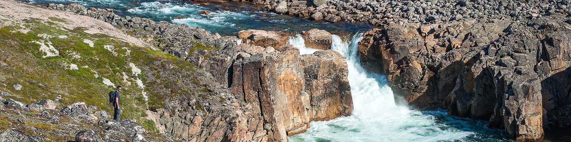 Un homme avec un sac à dos surplombant une cascade et un paysage de toundra rocheuse. 