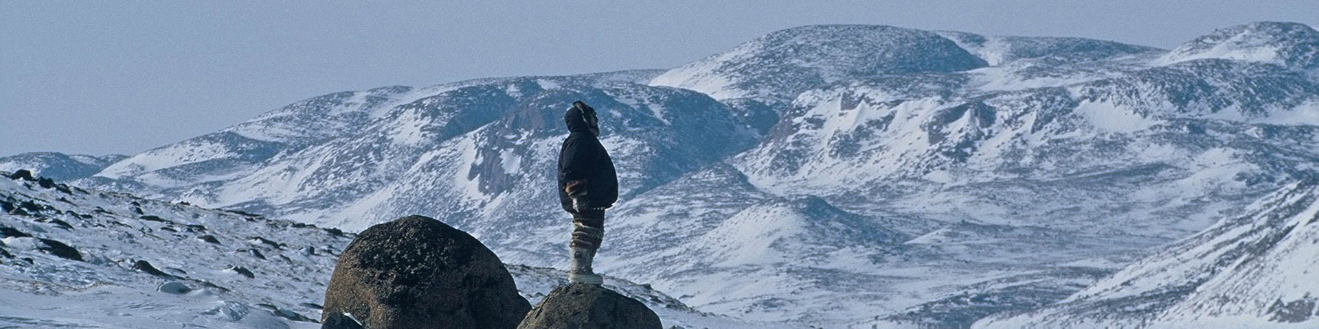 Un homme vêtu d'une parka se tient sur un gros rocher, entouré de montagnes et de paysages enneigés.