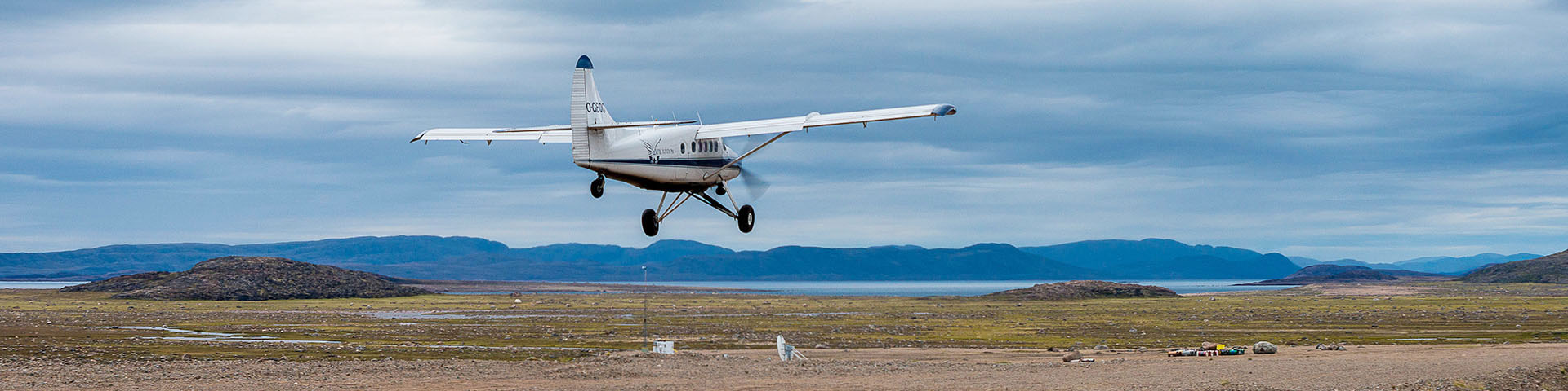 A small plane landing on a dirt runway with tundra scenery in the background.