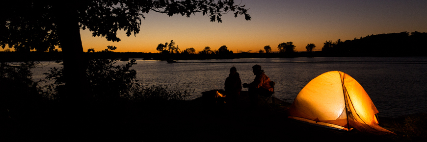 Two people sit beside a tent on the shoreline at sunset