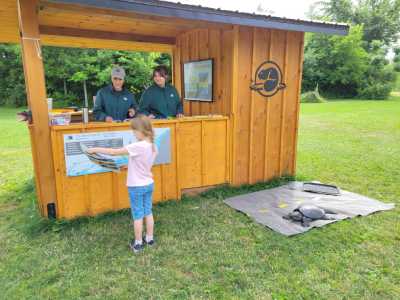 A child stands at a wooden park kiosk talking to two staff from inside.