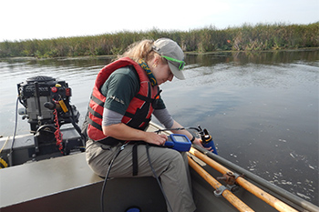 Un membre de l’équipe de conservation des ressources utilise un équipement spécialisé pour collecter des données sur la qualité de l’eau du marais. L’employé tient l’équipement hors d’un bateau à moteur et dans l’eau d’une main, tout en lisant les données sur un écran tenu dans l’autre. 
