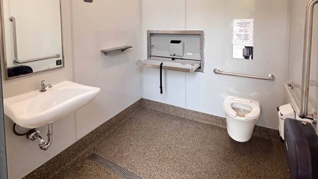 Une salle de toilette accessible équipée d’un lavabo, d’une toilette accessible, de barres d’appui et d’une table à langer rabattable.