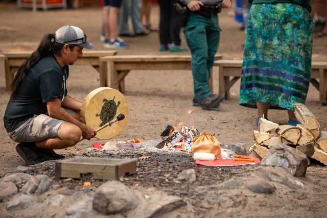 A person kneels beside a campfire while holding a hand drum during an outdoor cultural gathering,