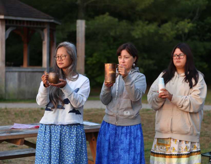 Three people stand outdoors holding small cups during a cultural activity, with trees in the background.