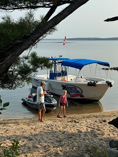 A boat is beached on a sandy shoreline next to a jet ski while an adult and child walk along the sand.