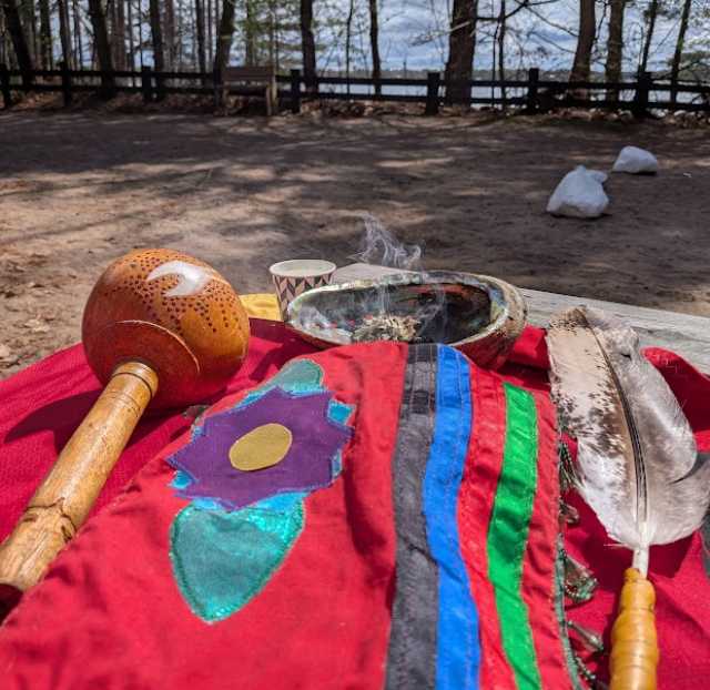 A drum, feather, and smudging bowl rest on a red cloth outdoors near the water