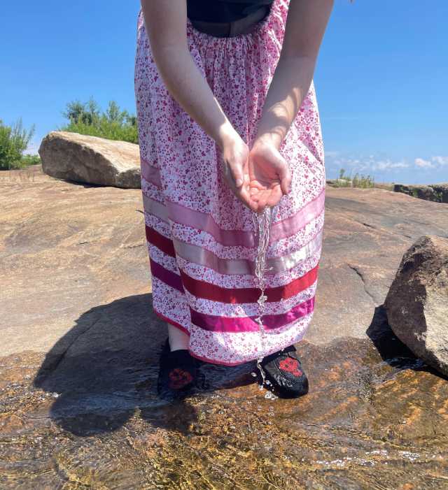 A person in a purple dress stands in shallow water, holding water in their hands.