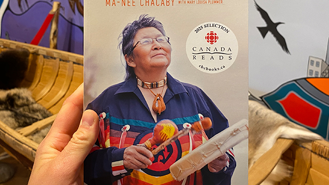 Une main tenant le livre intitulé « A Two-Spirit Journey » de Ma-Nee Chacaby. La couverture présente une femme autochtone en tenue traditionnelle, le regard tourné vers l’horizon, tenant un tambour.