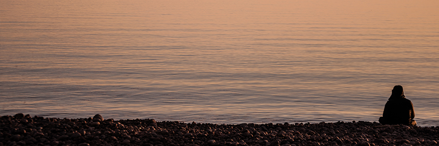 Person is sitting on a cobble beach looking out across the vast expanse of a calm Lake Superior at dusk