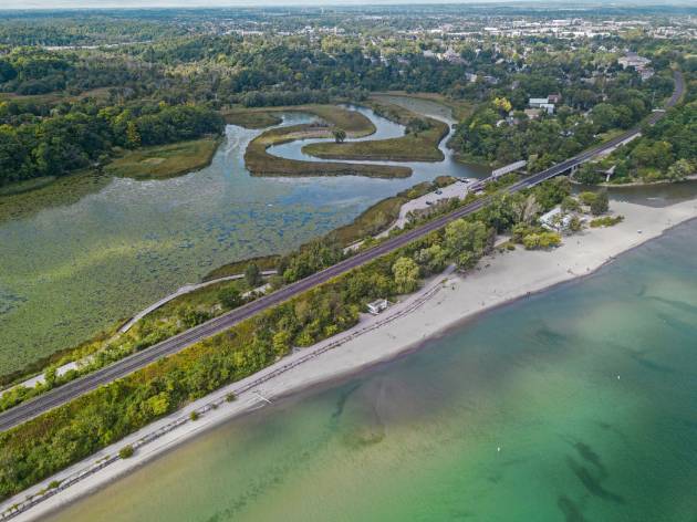 An aerial view of a rouge beach, a coastal landscape featuring a sandy beach with clear, greenish water. A railroad track runs parallel to the shoreline, separating the beach from a marshy area with a winding river. 