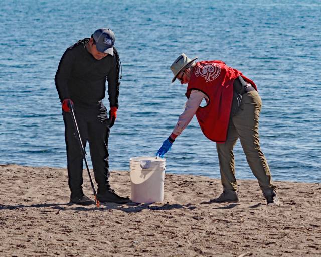 Deux personnes nettoient une plage. L'une, en noir avec une casquette, utilise un ramasseur de déchets, tandis que l'autre, en gilet et chapeau rouges, place les déchets dans un seau blanc. Le lac est à l'arrière-plan.