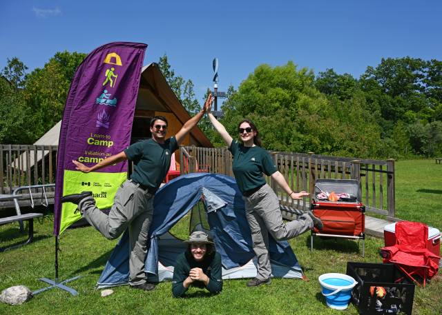 Un groupe de trois personnes est représenté dans une installation de camping, posant de manière ludique autour d'une petite tente. Deux d'entre eux se tiennent de chaque côté, en équilibre sur une jambe et se donnent la main, tandis que le troisième est allongé sur le sol, jetant un coup d'œil à l'extérieur de la tente. L'arrière-plan comprend une tente ressemblant à un tipi, un drapeau « Apprendre à camper » et divers équipements de camping tels qu'une glacière, des chaises et un réchaud portable.