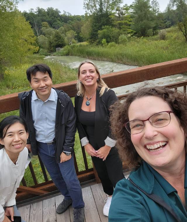 Quatre personnes se tiennent sur un pont en bois avec une rivière et de la verdure en arrière-plan. Elles sourient à apparel photo et prennent un selfie.